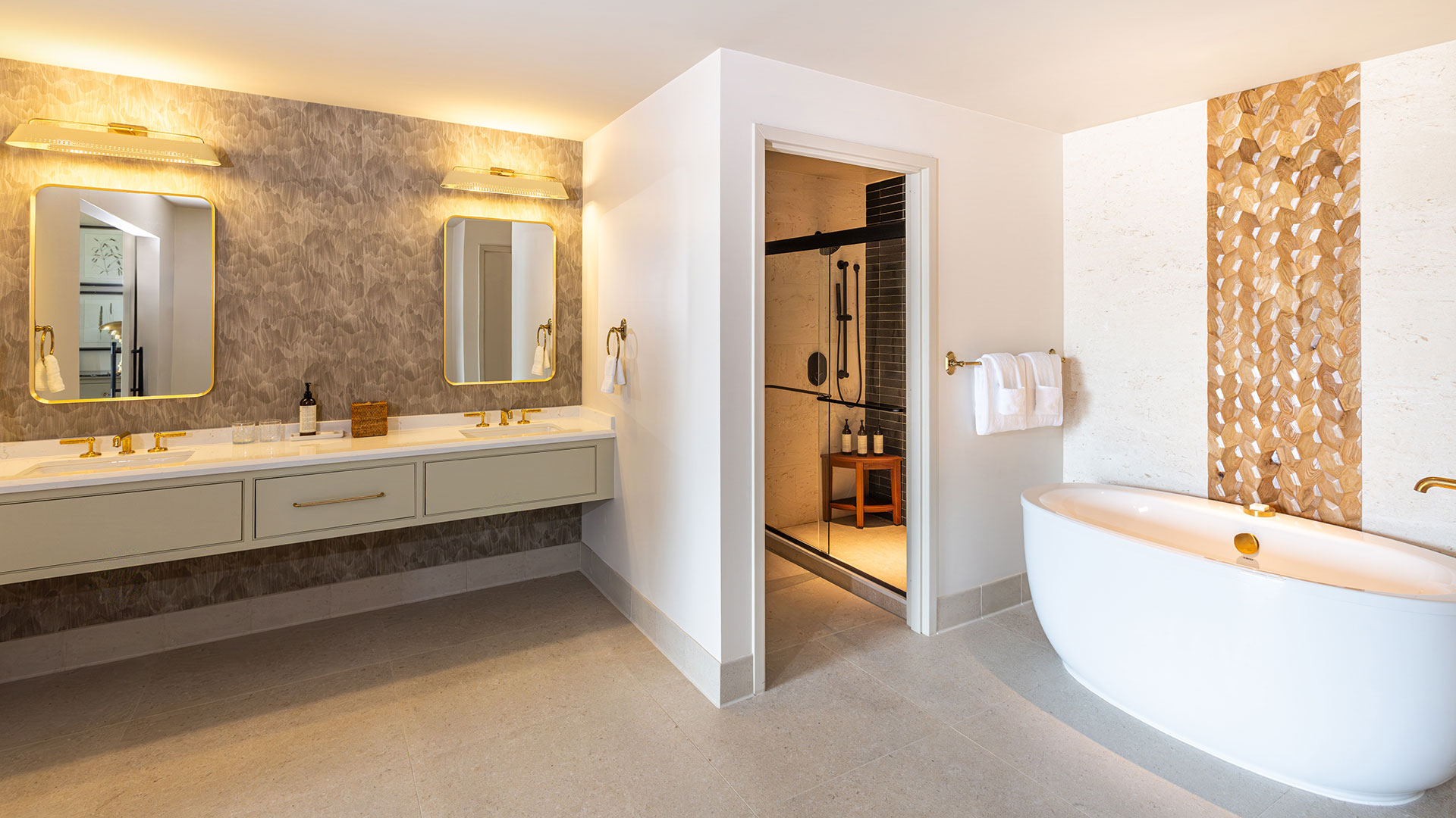 Spacious modern bathroom with double sinks and mirrors, a freestanding bathtub, gold accents, textured walls, and an open shower area with a glass door. Towels and toiletries are neatly arranged.