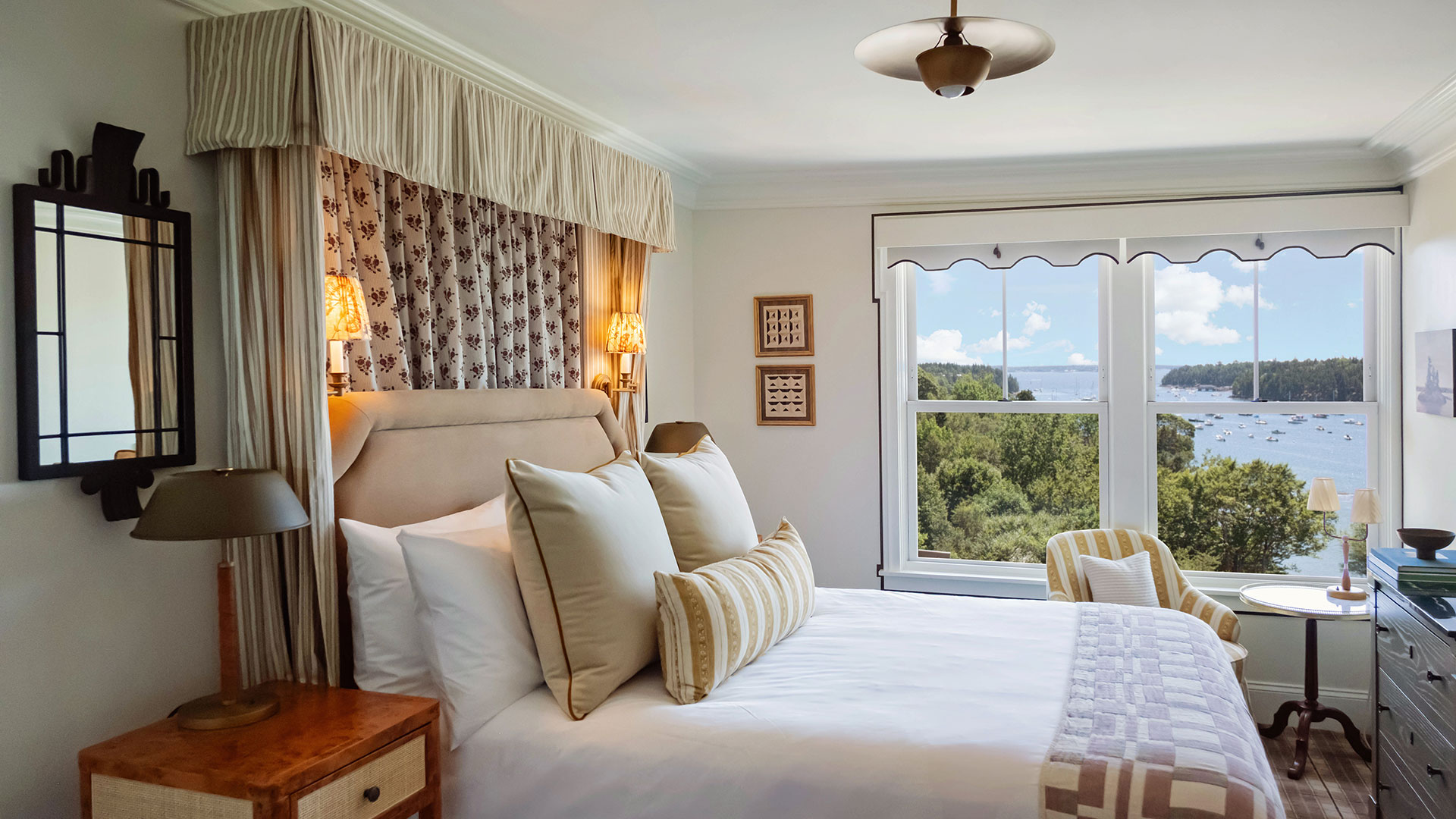 A cozy, well-lit bedroom with a large bed, elegant decor, lamps, a window overlooking trees, water, and boats, and a small table with chairs near the window.