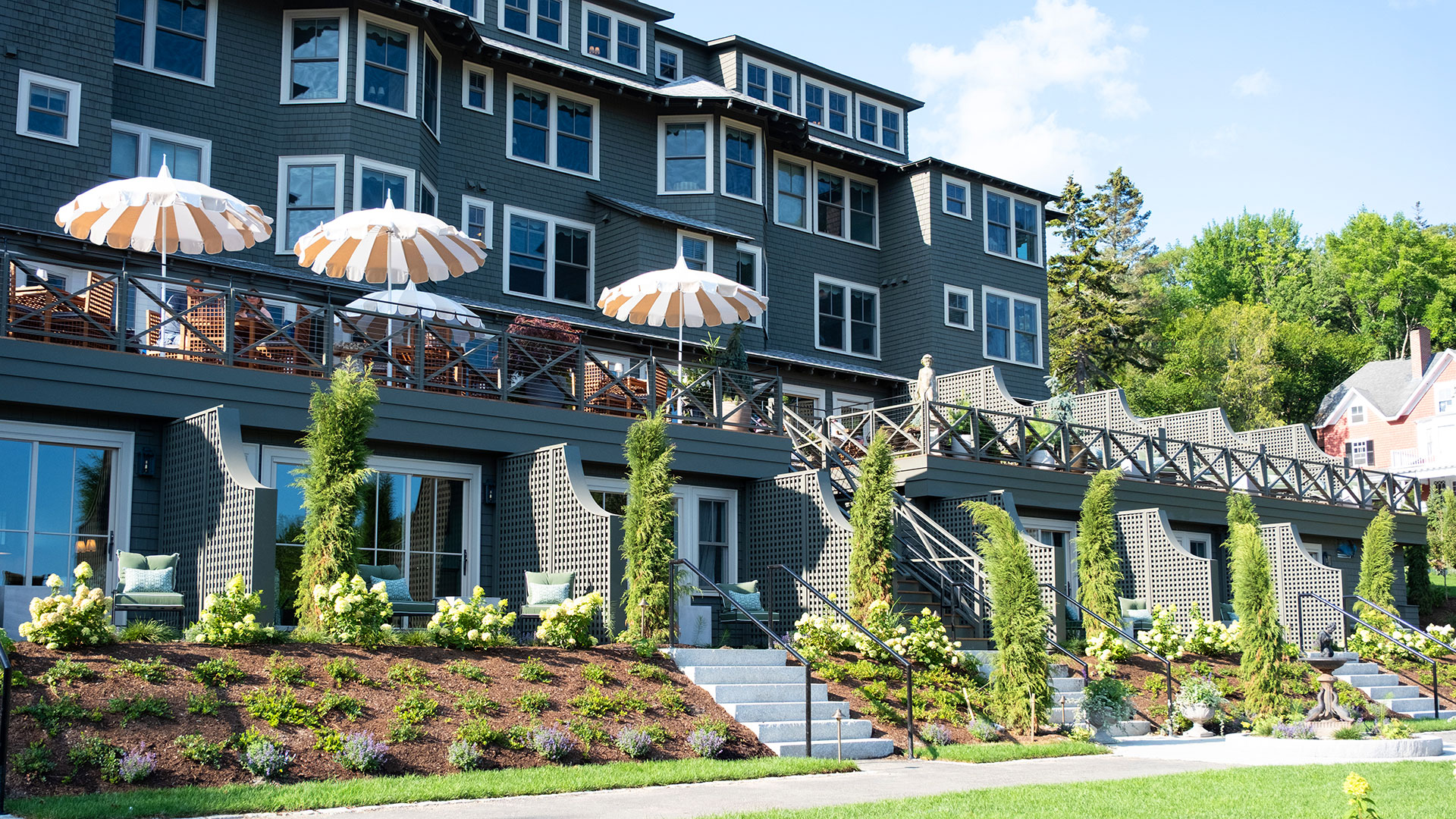 A large gray hotel with striped umbrellas on a balcony, greenery, flowering plants, and steps leading up the landscaped hill under a blue sky.