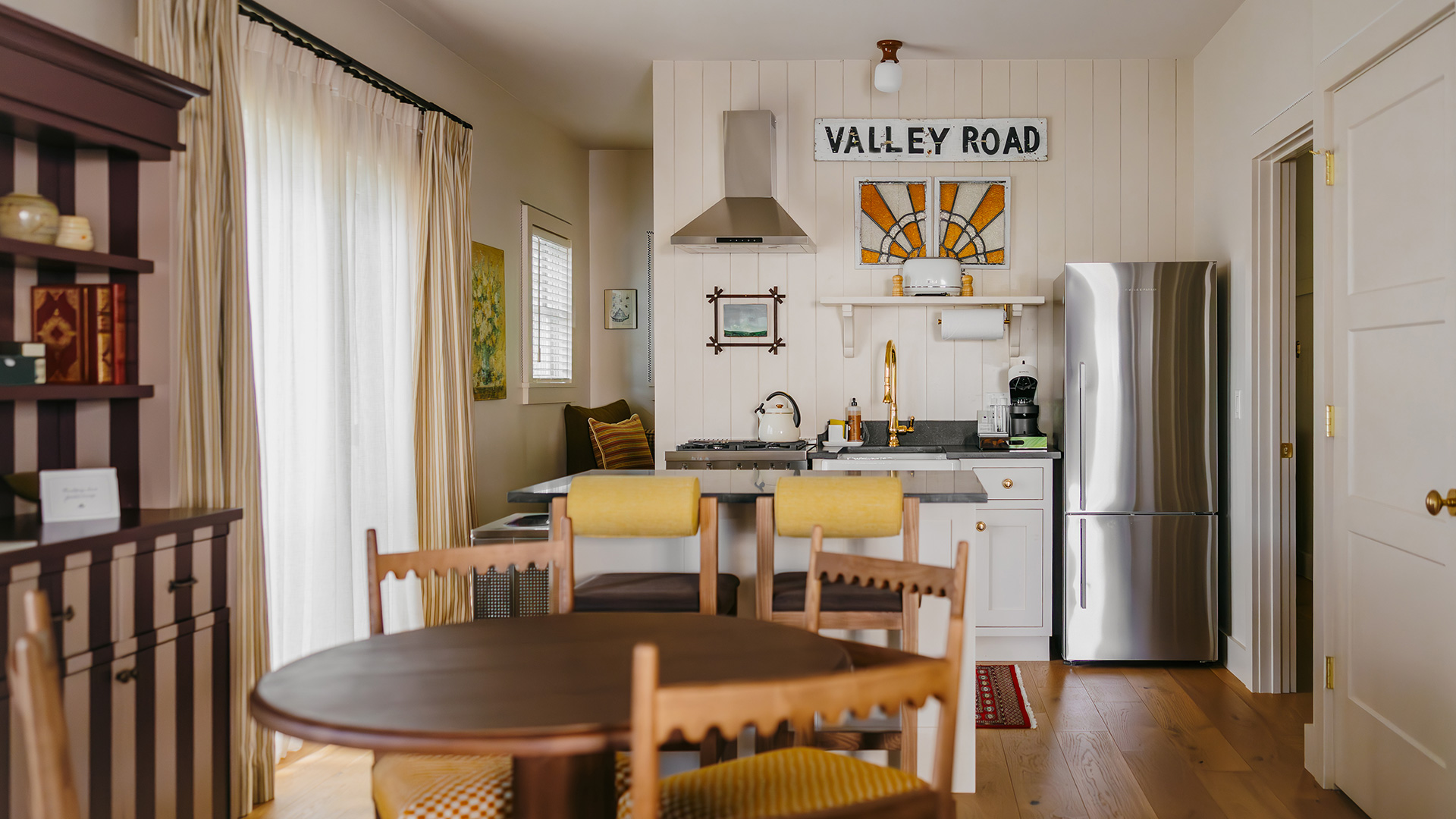 Small, cozy kitchen with a round wooden dining table, yellow cushioned chairs, stainless steel fridge, stove, and Valley Road sign above the sink. Sunlight filters through sheer curtains on the left.