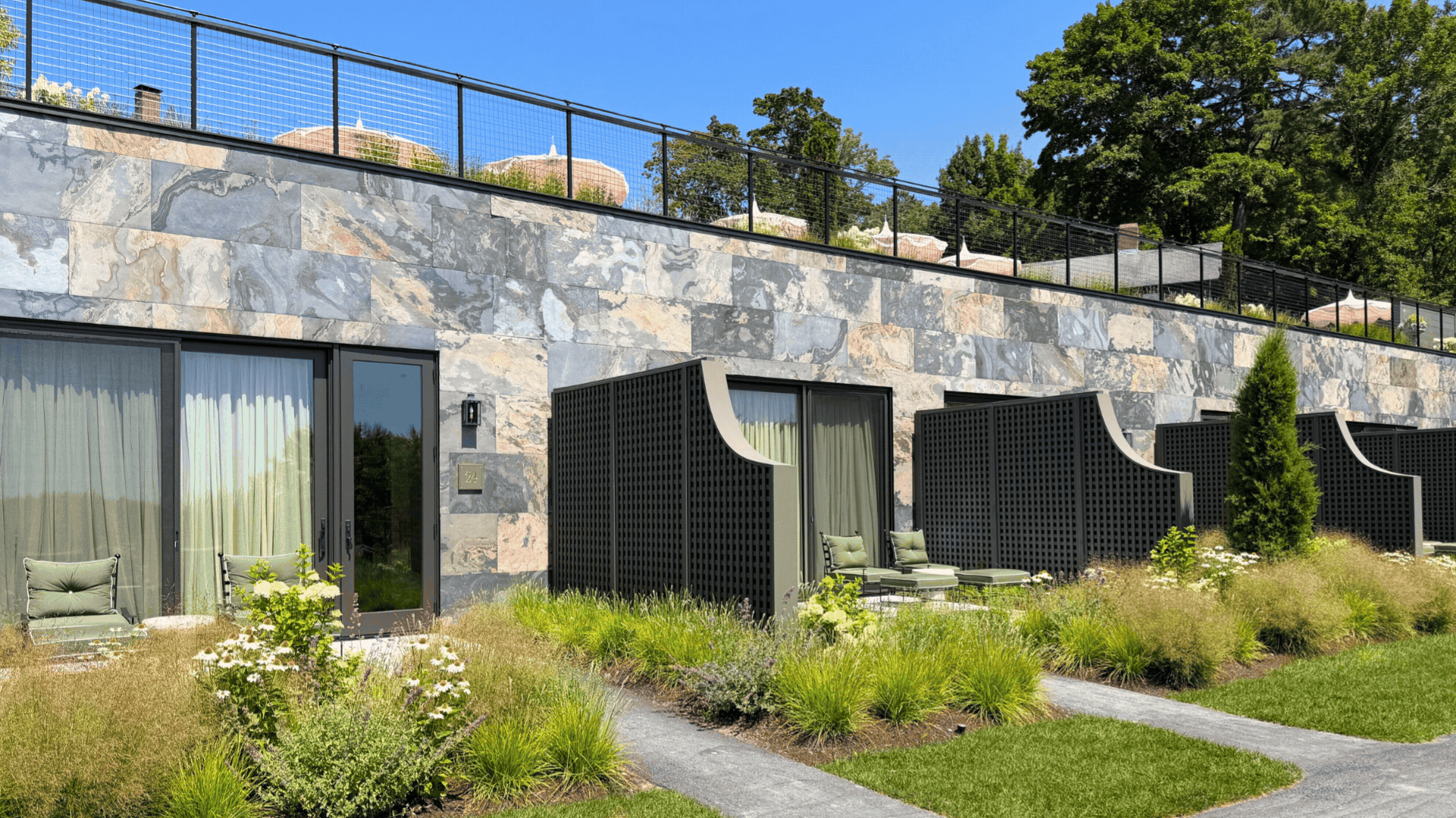 Modern stone building with private patios, green chairs, and black privacy screens, surrounded by landscaped greenery, under a clear blue sky.