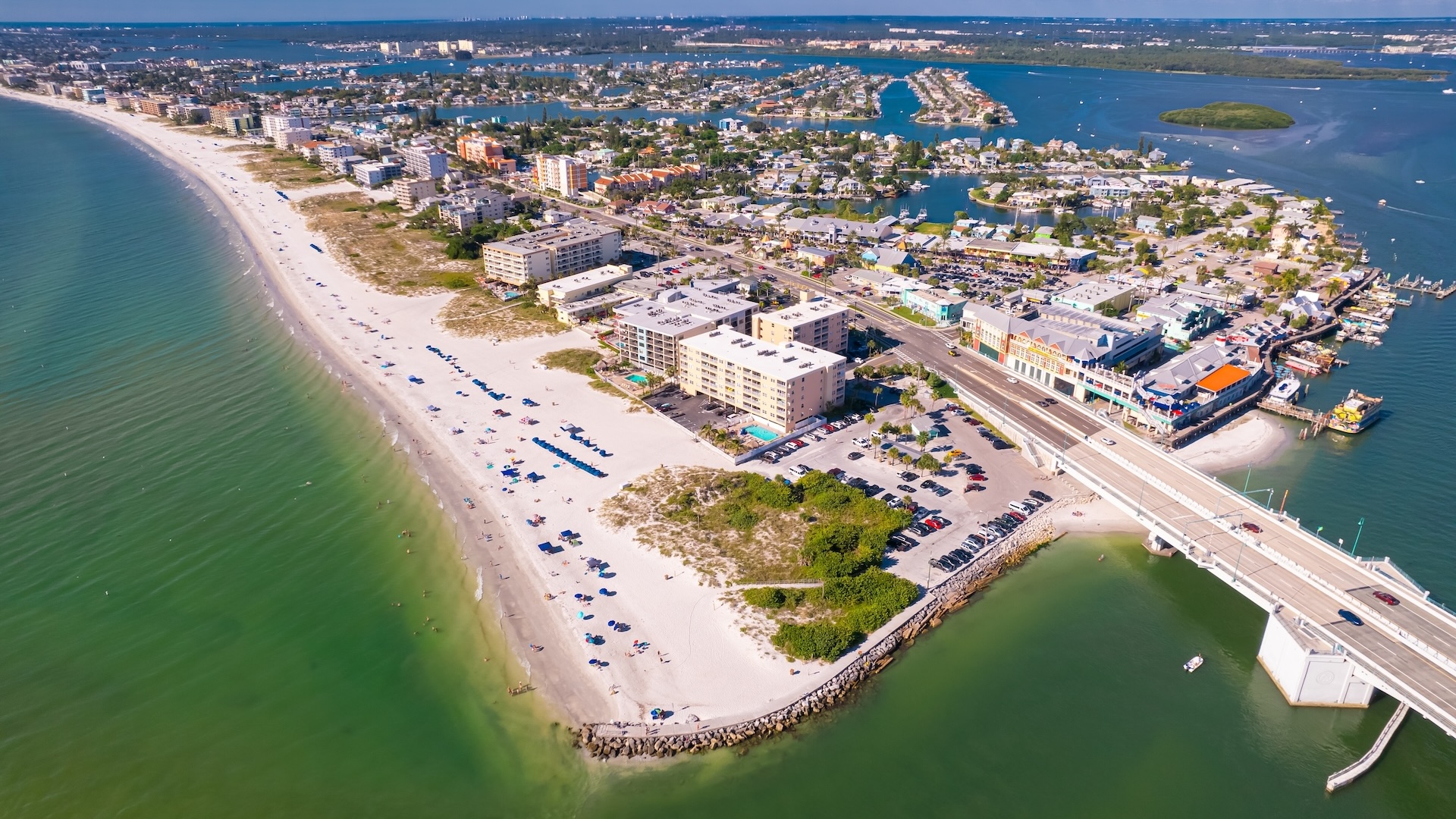 Aerial view of a coastal town with sandy beaches, ocean waves, buildings, parked cars, and a bridge connecting the shoreline to the city.