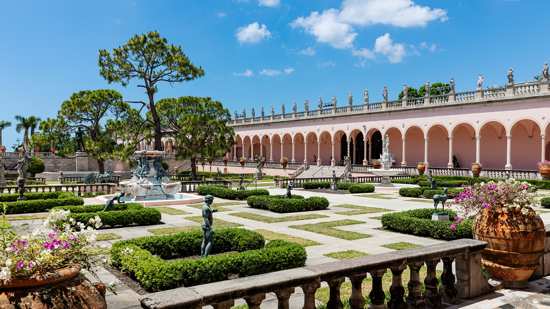 A formal garden with manicured hedges, statues, a central fountain, and a long pink building with arches and sculptures on the roof under a sunny blue sky.