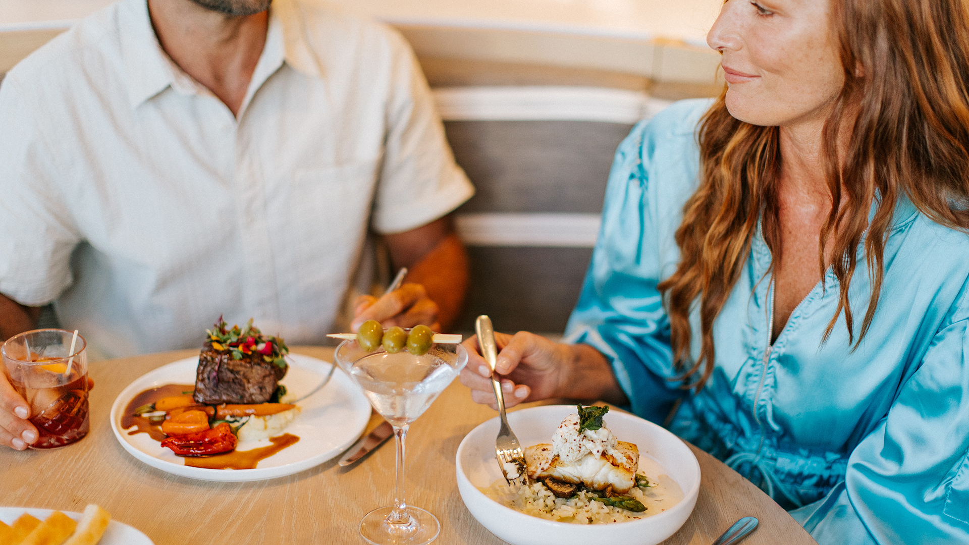 A woman in a blue blouse and a man in a light shirt sit at a table sharing a meal, with plates of food and cocktails in front of them, smiling and enjoying their time together.