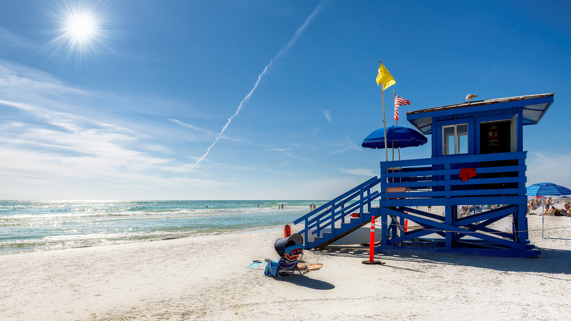 A bright, sunny day at the beach with a blue lifeguard tower, yellow flag, people along the shoreline, and a clear sky with a visible sun and contrail.