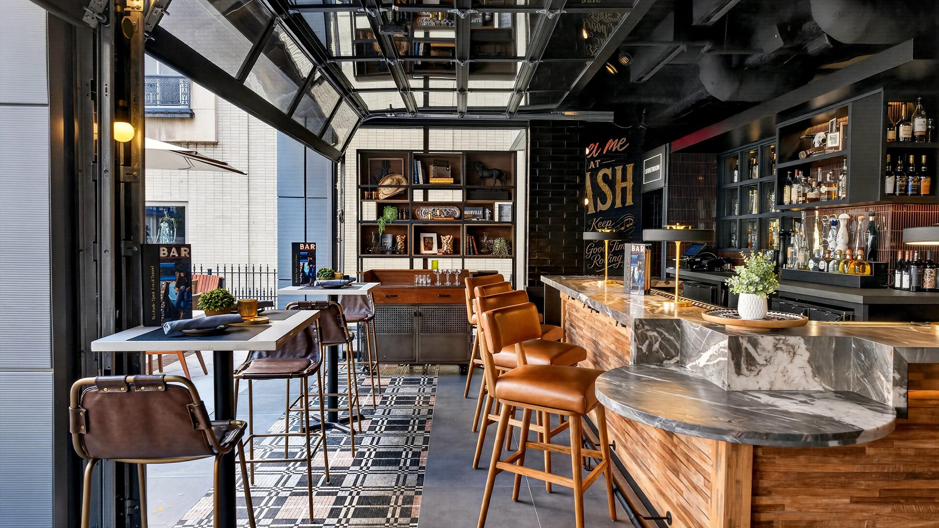 Modern bar with wooden and marble accents, brown leather barstools, open glass doors, shelves with bottles and decor, and stylish tile flooring. Some tables and chairs are set near the entrance.