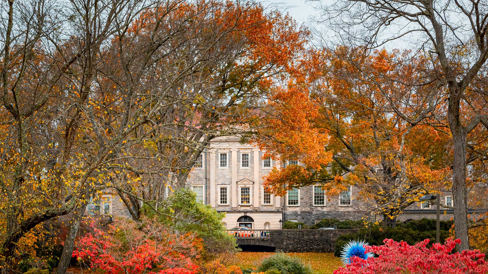 A large, historic building is partially hidden by trees with orange and yellow autumn leaves, with red foliage and a blue sculpture in the foreground.