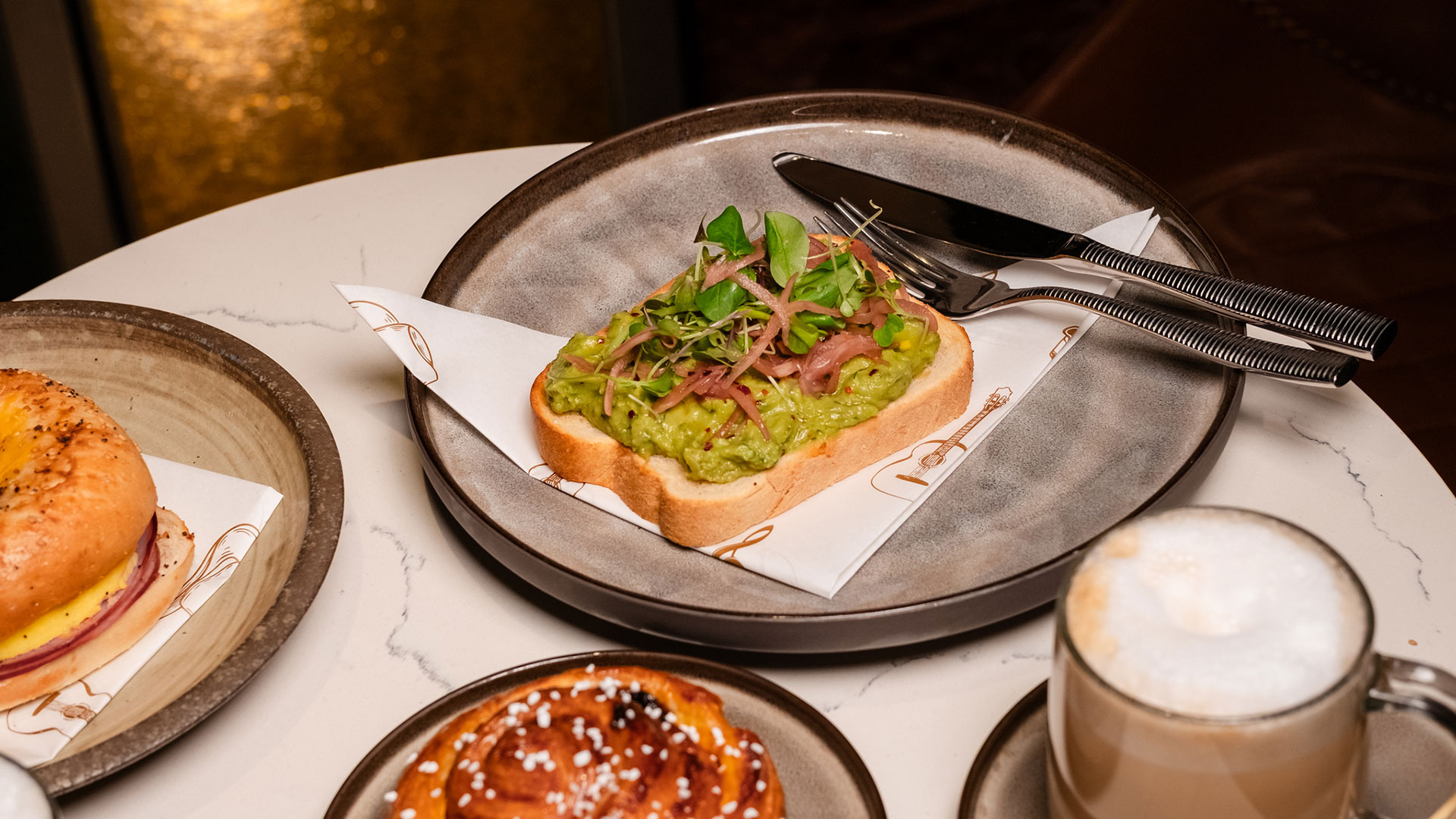 A plate with avocado toast topped with greens and onions, placed on a napkin with a knife and fork beside it. Nearby are a bagel sandwich, a pastry, and a cup of cappuccino on a marble table.