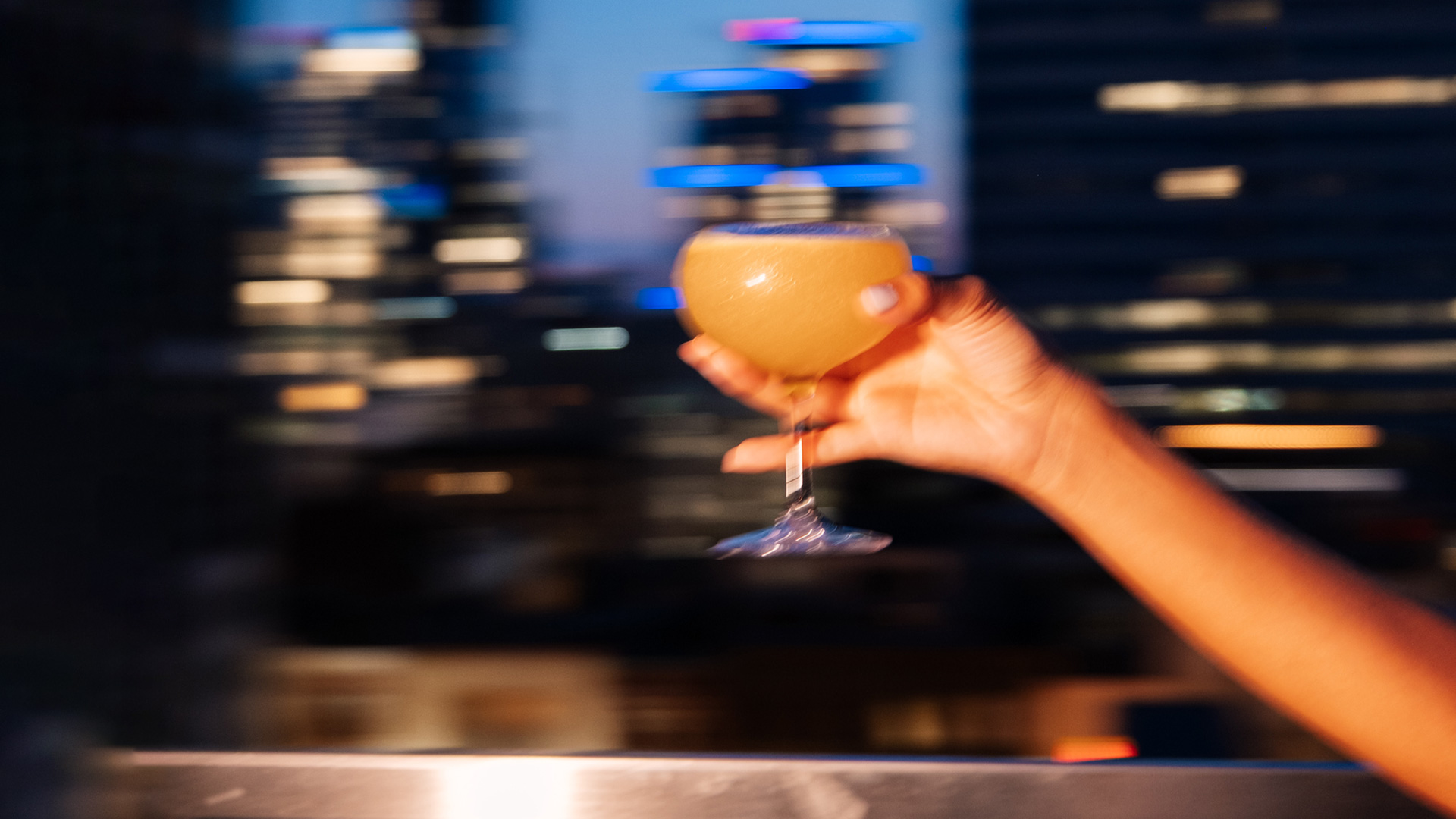 A hand holds a cocktail glass with an orange drink, against a blurred cityscape backdrop featuring colorful lights and tall buildings at dusk.