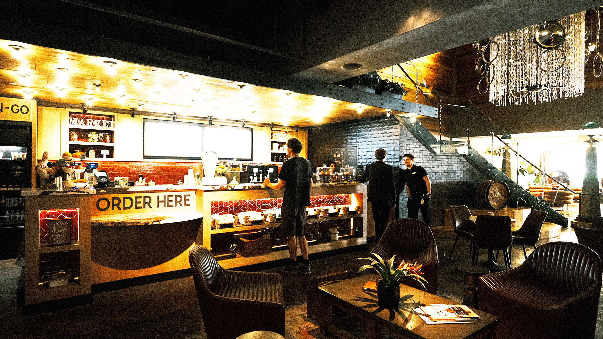 A brightly lit café counter with a large Order Here sign. Several people are ordering, while others stand nearby. Cozy seating, tables, and modern décor fill the foreground, with a staircase visible in the background.