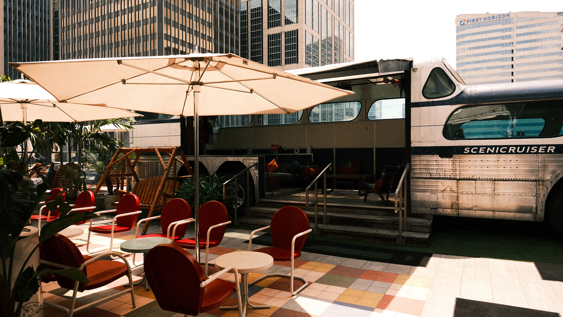 Outdoor seating area with red chairs and white tables under large umbrellas, next to a retro silver bus with an open side in a city setting with tall office buildings in the background.