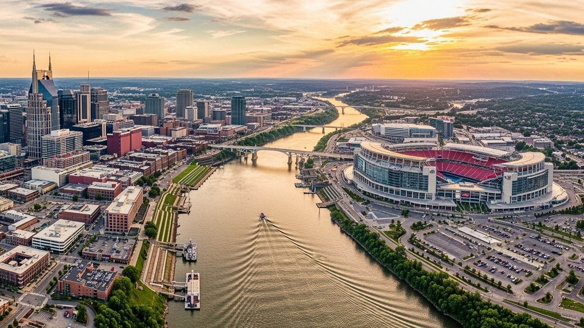 Aerial view of downtown Nashville at sunset, showing a river, city buildings, a bridge, and a large stadium with parking lots, all under a colorful sky.