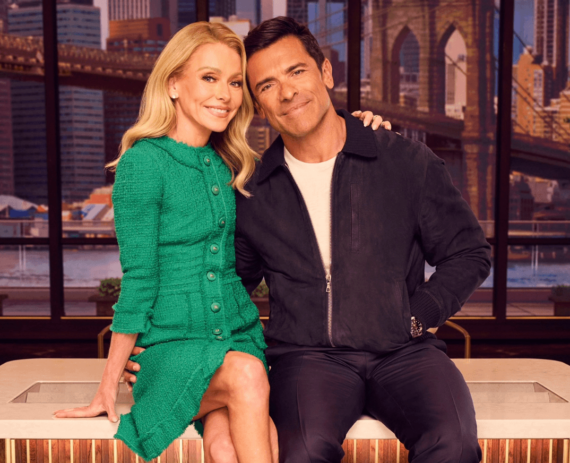 Kelly Ripa and Mark Consuelos sitting together in a television studio with a bridge visible through the window.