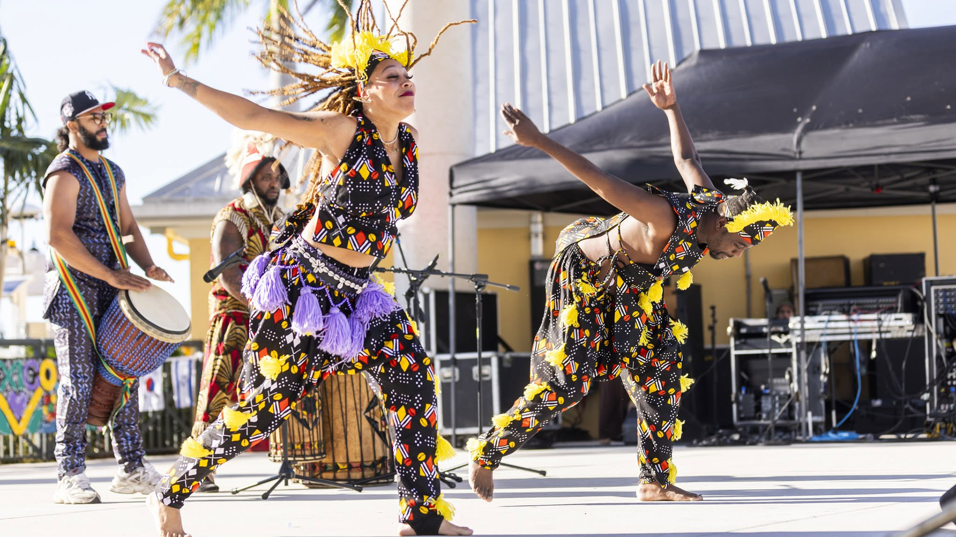 Two dancers in vibrant, patterned outfits with yellow hair accessories perform on an outdoor stage, accompanied by a drummer.