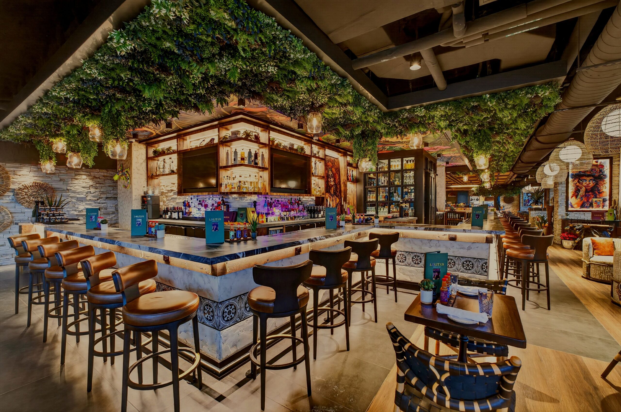 An interior view of a bar and restaurant featuring a central stone bar with leather stools, hanging greenery from the ceiling, and several small dining tables.