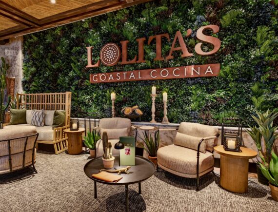 Lounge area at Lolitas Coastal Cocina with armchairs and a sofa set against a large indoor wall of greenery featuring the restaurants logo.