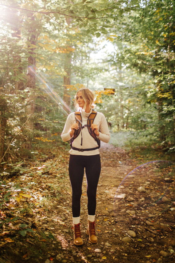 A woman with a backpack stands on a forest trail, surrounded by tall green trees. She is wearing a light jacket, black leggings, and boots, with sunlight streaming through the leaves.
