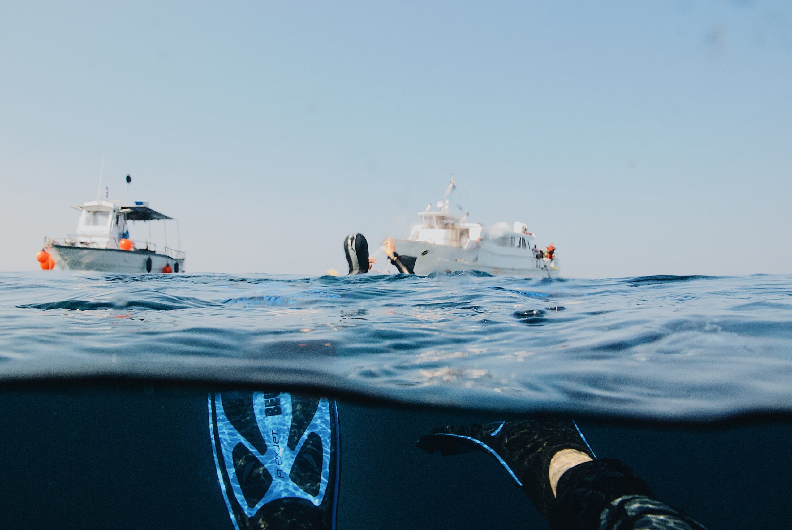A person snorkeling in the ocean, half submerged in water, with dive fins visible. Two boats float on the horizon in the background under a clear blue sky.