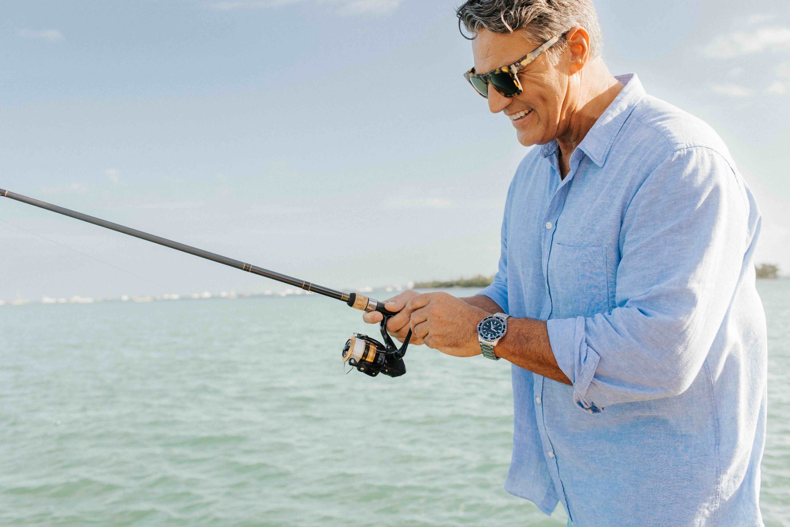 A man in a light blue shirt and sunglasses is smiling while holding a fishing rod. He stands on a boat surrounded by water under a clear blue sky.