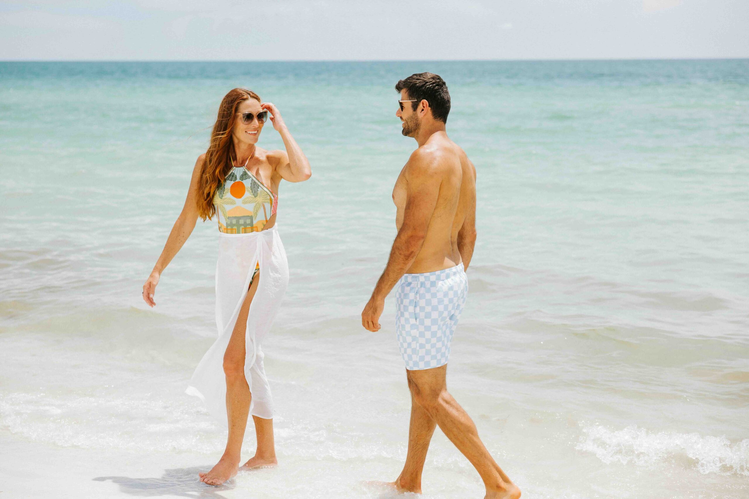 A woman in a colorful swimsuit with a white wrap and a man in checkered swim trunks walk along the ocean shore. The sky is clear and the sea is calm.