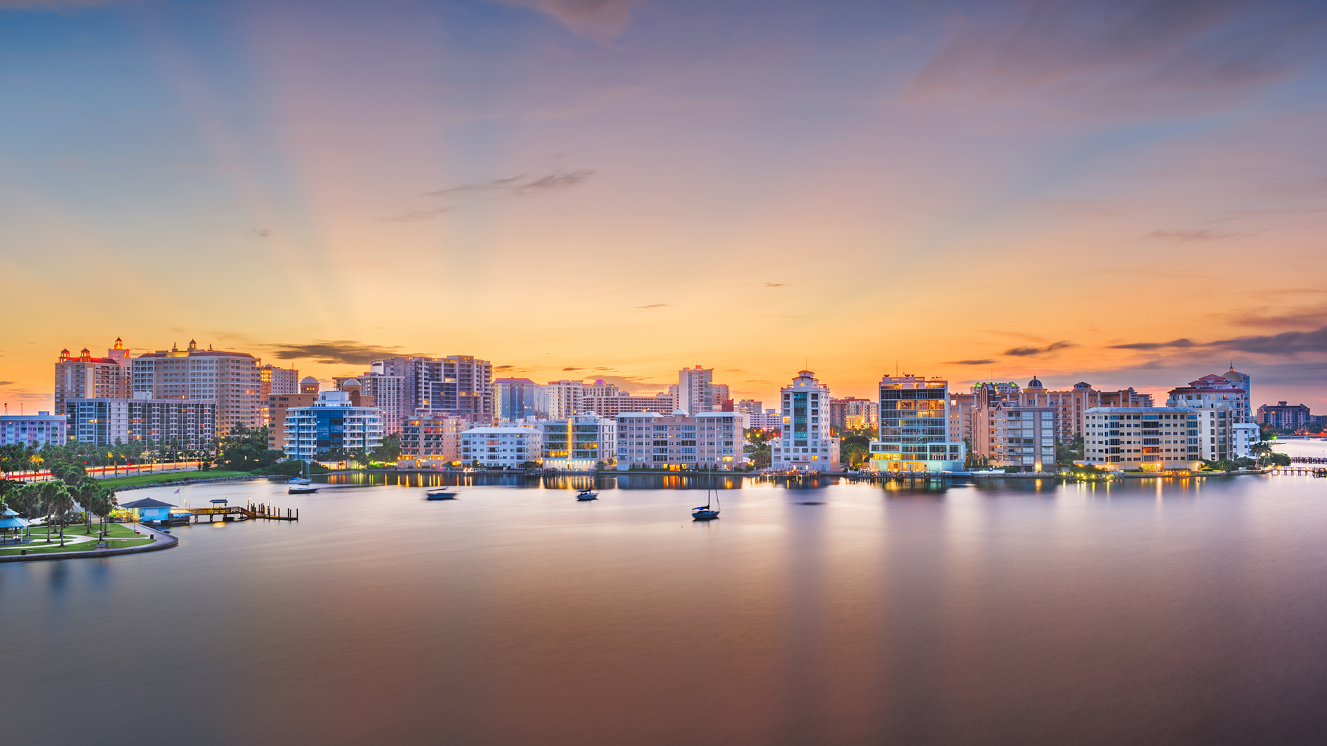 Skyline view of a coastal city at sunset, with tall buildings reflecting in the calm water. The sky displays a gradient of orange, yellow, and blue hues, with a few small boats visible in the foreground.