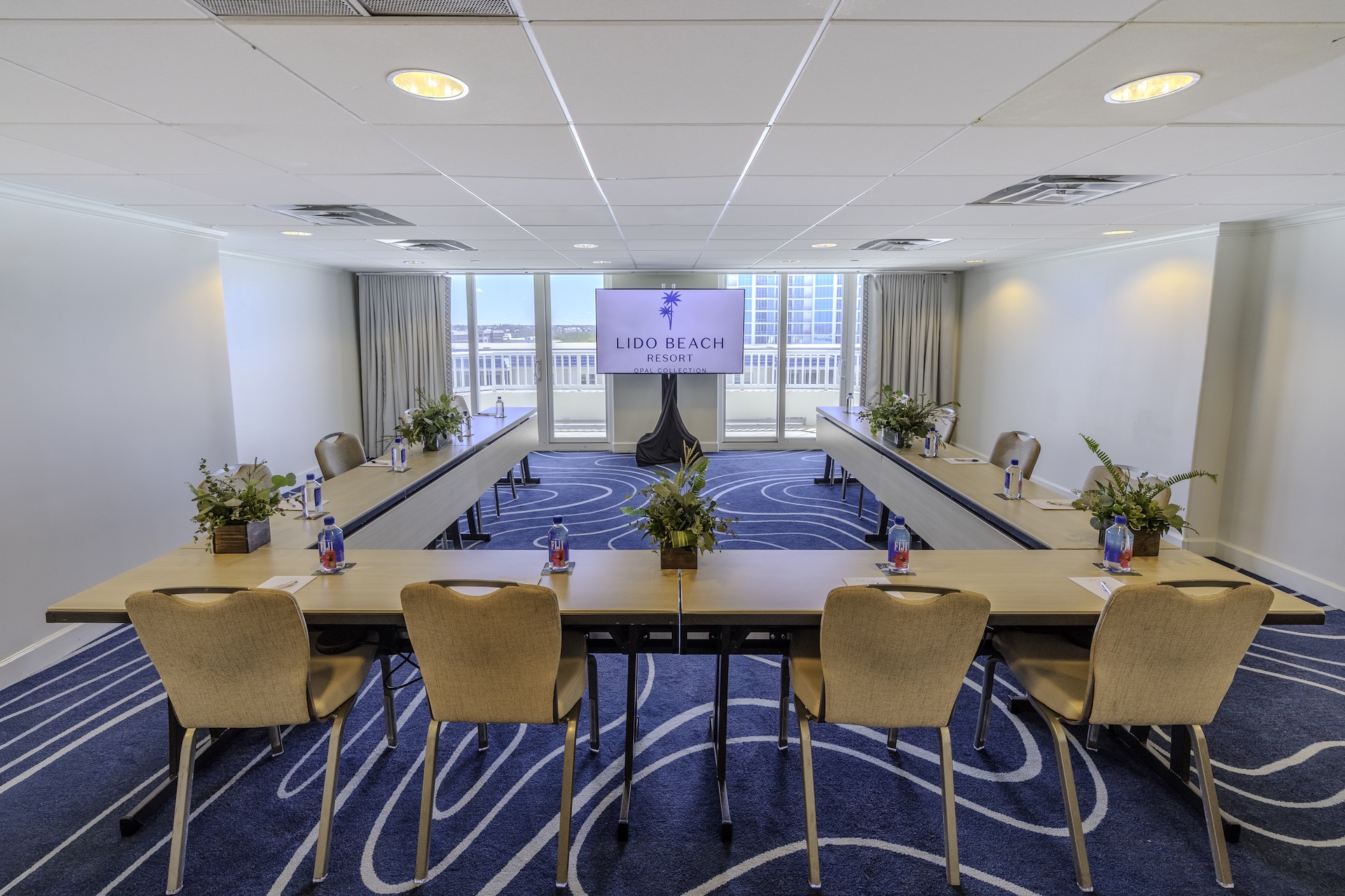 A modern conference room with U-shaped tables, beige chairs, flower centerpieces, and bottled water, facing a screen displaying Lido Beach Resort. Large windows at the back show a view of the outdoors.