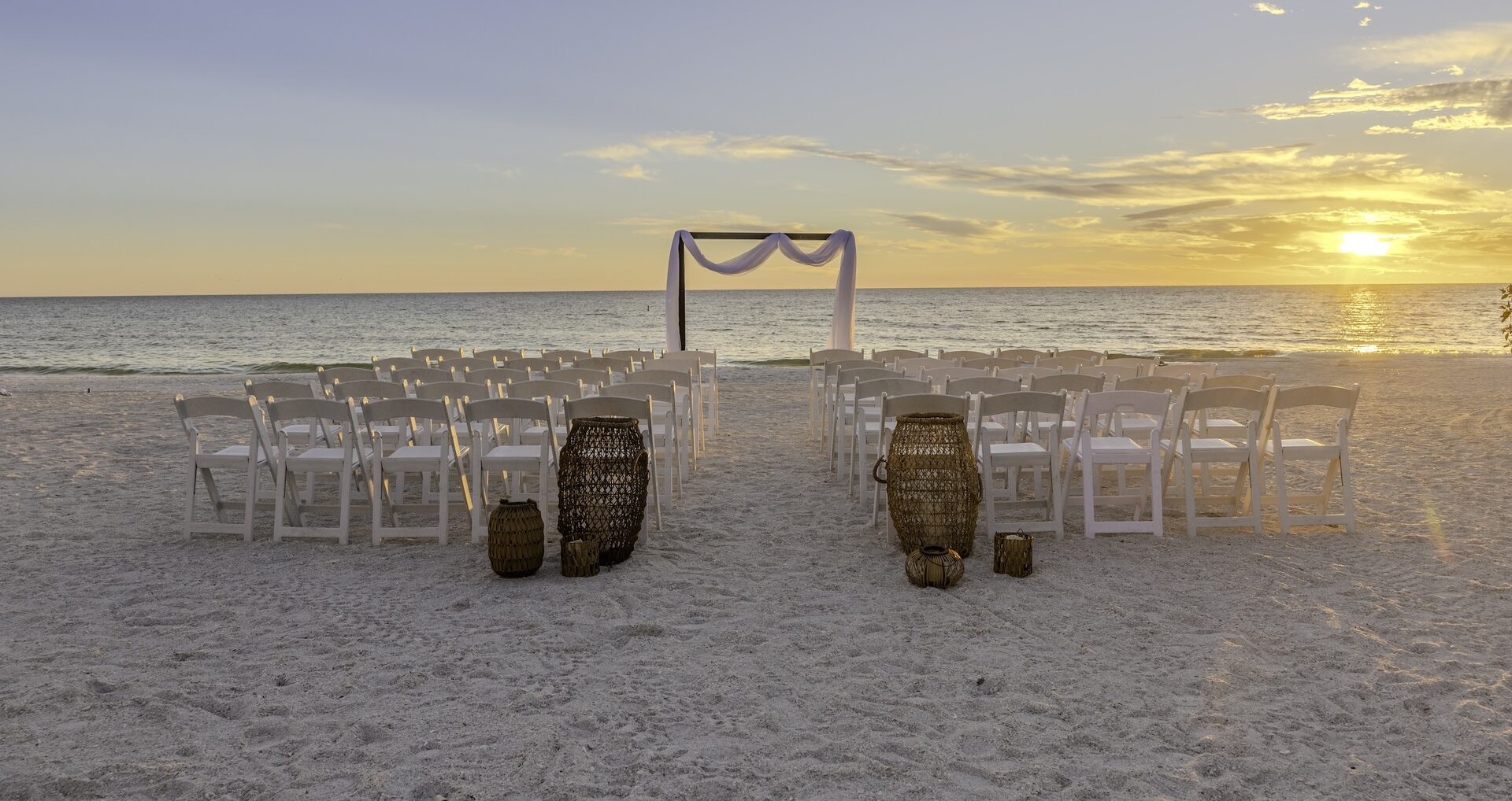 Rows of white chairs face a simple wedding arch draped with white fabric on a sandy beach at sunset, with decorative wicker baskets in the foreground and the ocean in the background.