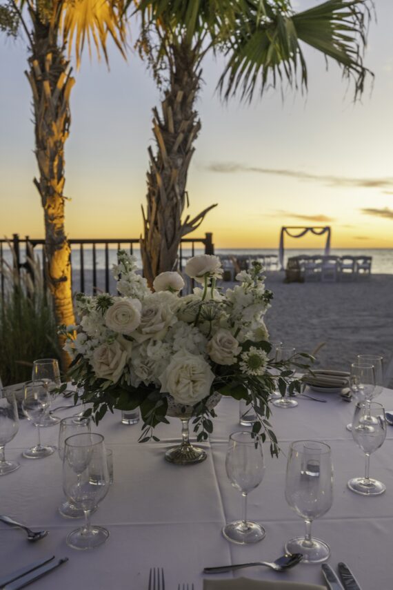 A round table set for a formal event with a centerpiece of white flowers, surrounded by glassware, at a beach during sunset. Palm trees and an archway on the sand are visible in the background.