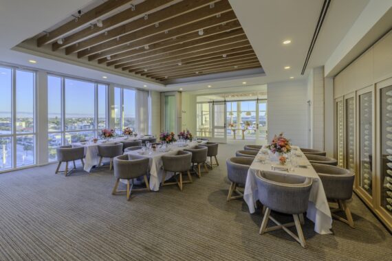 A modern dining room with large windows, wooden ceiling beams, and neatly set tables with gray chairs and colorful flower centerpieces, overlooking a scenic outdoor view.