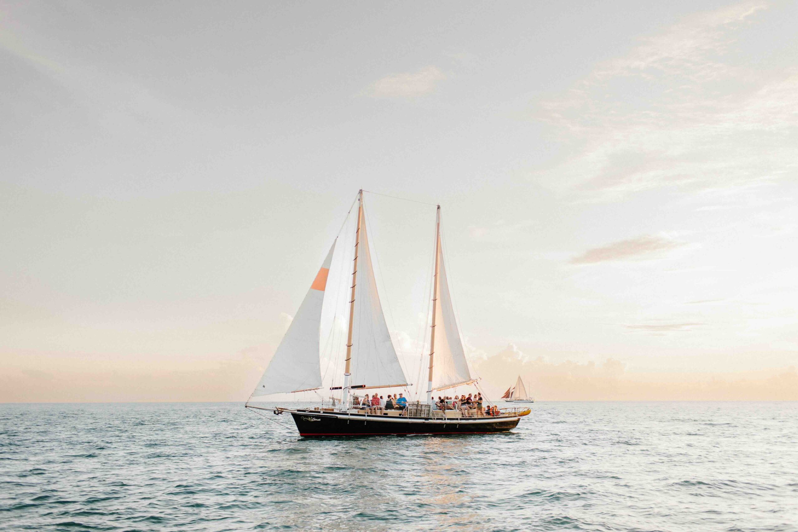 A sailboat with two white sails glides across calm ocean water at sunset, carrying several people on board. The sky is mostly clear with a soft golden glow.