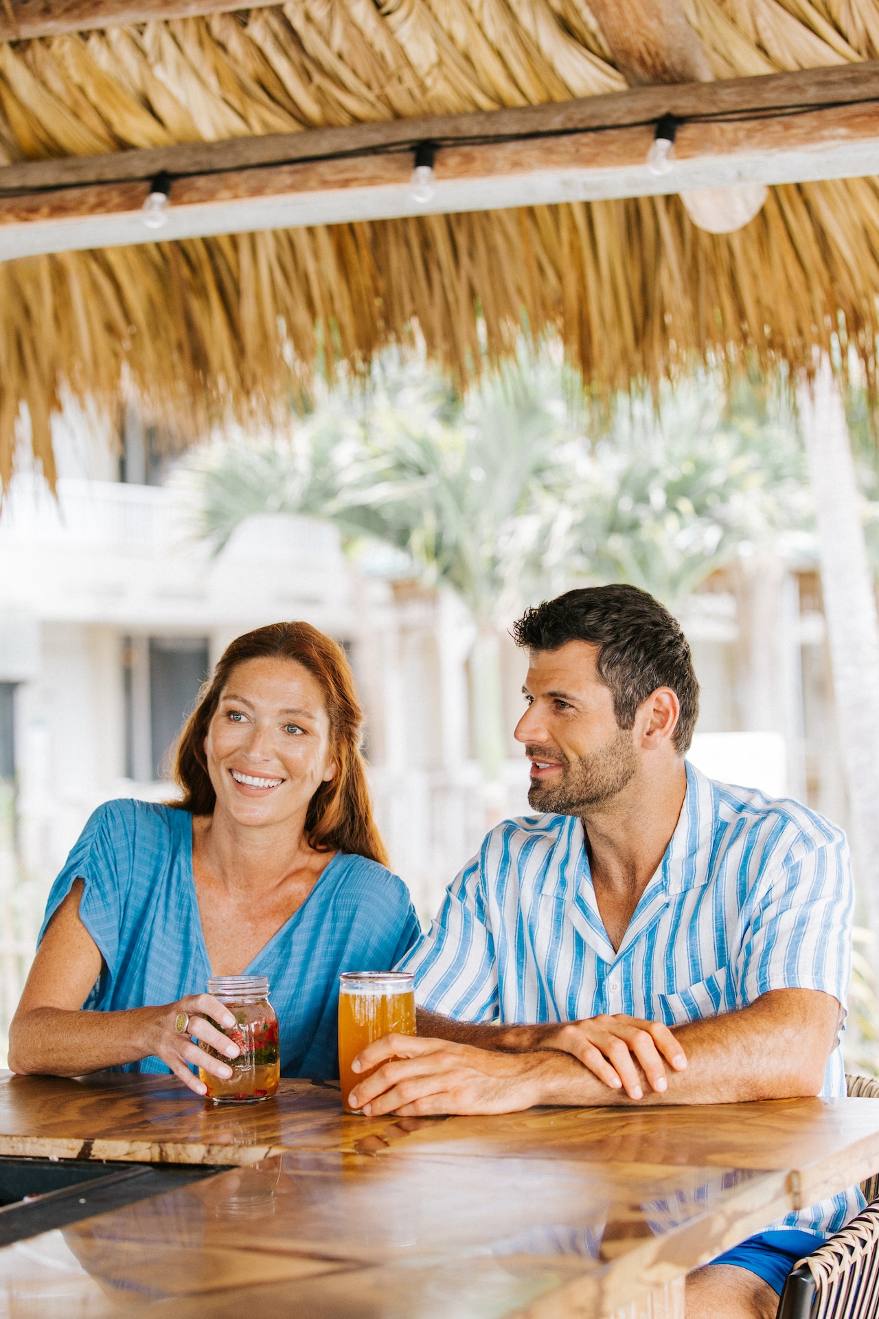 A smiling woman and man sit at a wooden bar under a thatched roof, each holding a drink, with tropical trees and buildings visible in the background.