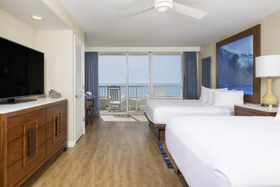 Hotel room with two beds, a wooden dresser, a television, and a balcony overlooking the ocean.