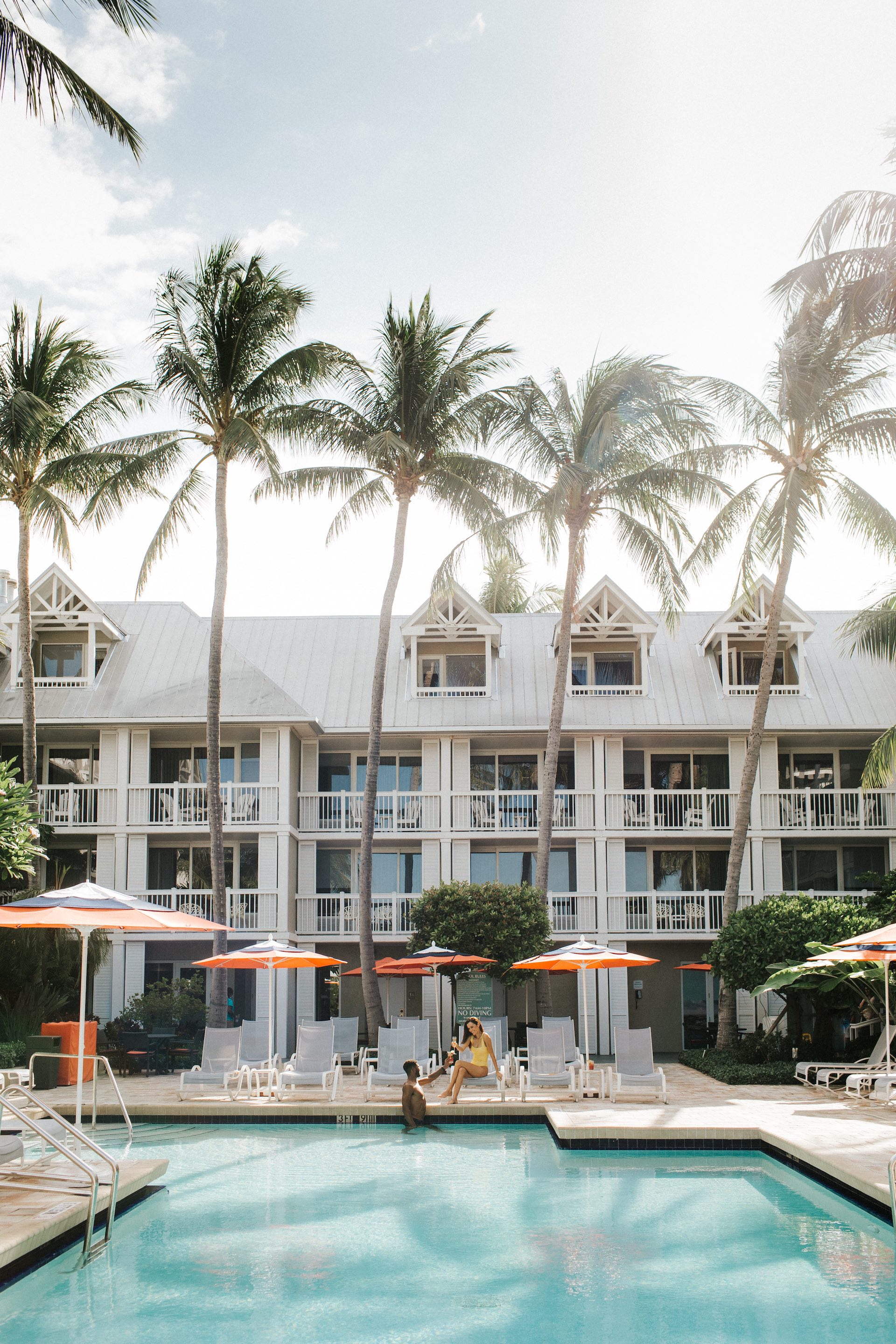 A large white building with balconies and a pool in front of it, surrounded by palm trees and lounge chairs.