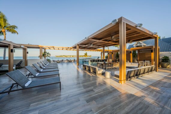 Outdoor poolside lounge area with modern wooden pergolas, sun loungers, and a seating area overlooking the ocean under a clear sky. The space has palm trees and elegant, contemporary décor.