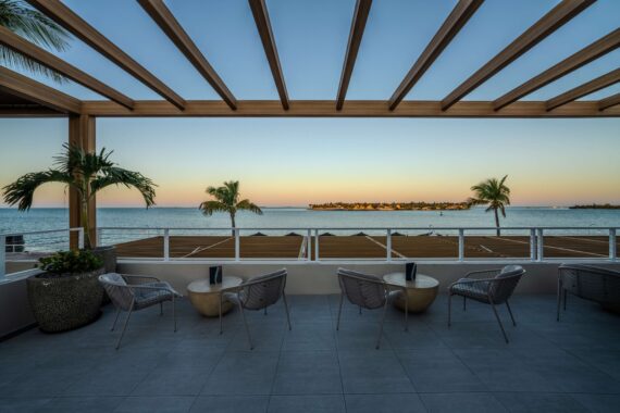 Modern terrace with chairs and small tables overlooks the ocean at sunset, framed by wooden pergola beams above and palm trees along the shore. The sky is clear, and the water is calm and blue.