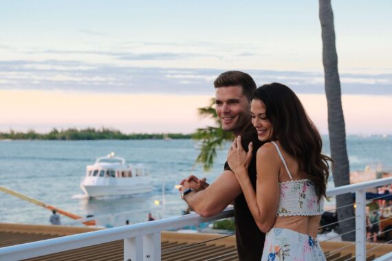 A smiling couple stands by a waterfront railing at sunset, with a boat on the water and palm trees in the background. The woman hugs the man from behind, both appearing happy and relaxed.