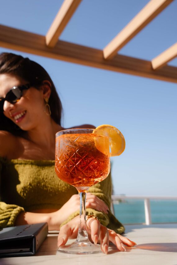 A woman wearing sunglasses and a green top smiles at an outdoor table with a glass of orange-colored cocktail garnished with a lemon slice, set against a sunny, blue sky and ocean backdrop.