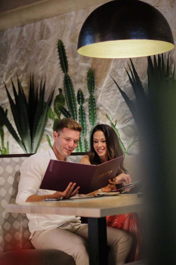 A man and woman sit together at a restaurant booth, smiling as they look at a menu. Large green cacti and plants are in the background, and a round black lamp hangs above their table.