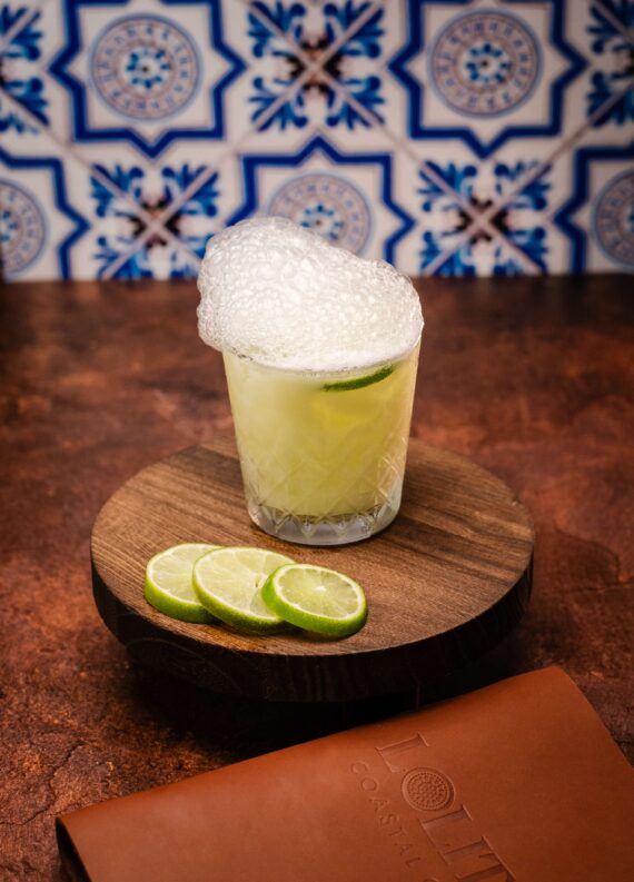 A cocktail in a glass with frothy foam on top and a lime slice inside, sits on a round wooden board with three lime slices beside it. The background features blue and white patterned tiles.