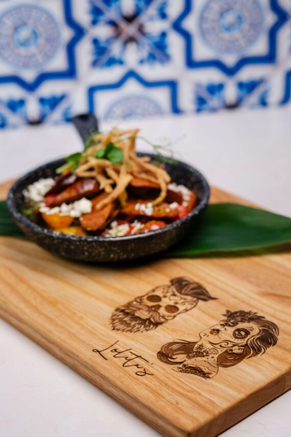 A skillet of food garnished with herbs and crispy strips sits on a wooden board decorated with Day of the Dead-style skull illustrations and the word Lottos, with blue and white tile in the background.