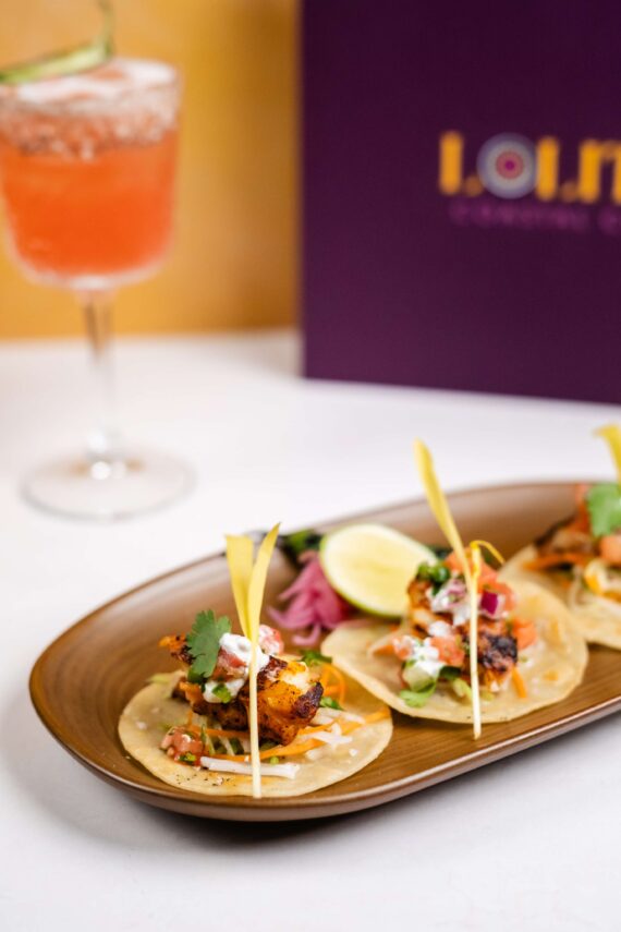 A plate with three small tacos garnished with cilantro, pickled onions, and lime wedge sits on a table. A pink cocktail in a glass and a purple menu are in the background.