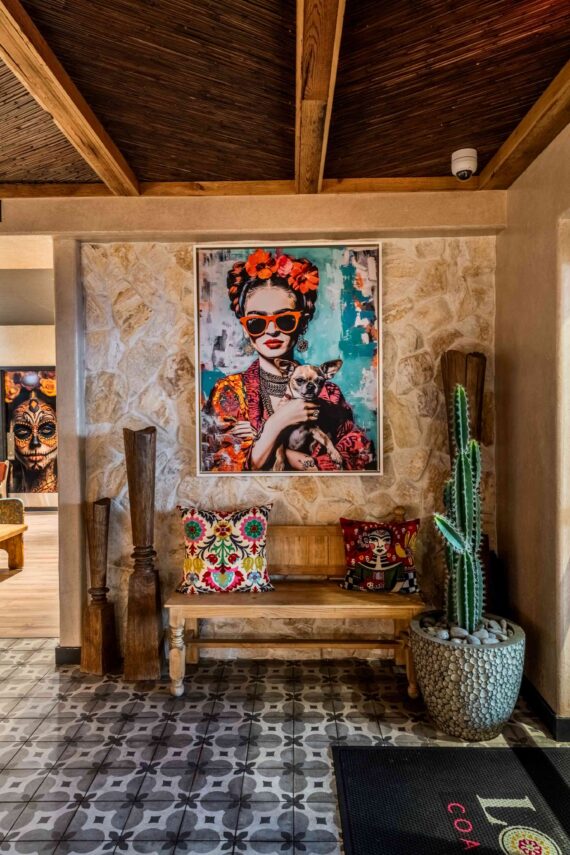 A vibrant, colorful portrait of a woman with flowers in her hair and sunglasses hangs above a wooden bench with decorative pillows, next to a tall cactus and carved wooden pieces in a stylish, rustic room with patterned tile flooring.