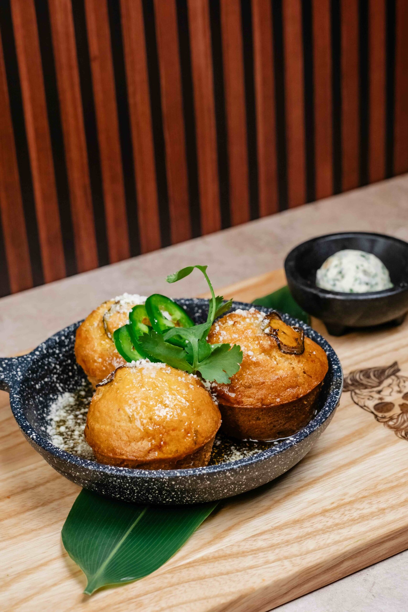 Three golden cornbread muffins topped with grated cheese, jalapeño slices, and cilantro are served in a black pan on a wooden board. A small bowl of herbed butter is placed in the background.
