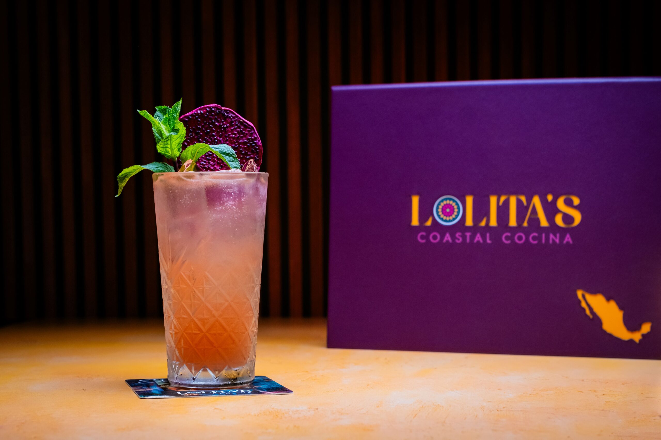 A tall, frosted cocktail glass with a pink drink, garnished with mint leaves and a purple slice, placed on a coaster. In the background, a purple menu titled Lolitas Coastal Cocina with a Mexico map outline is visible.