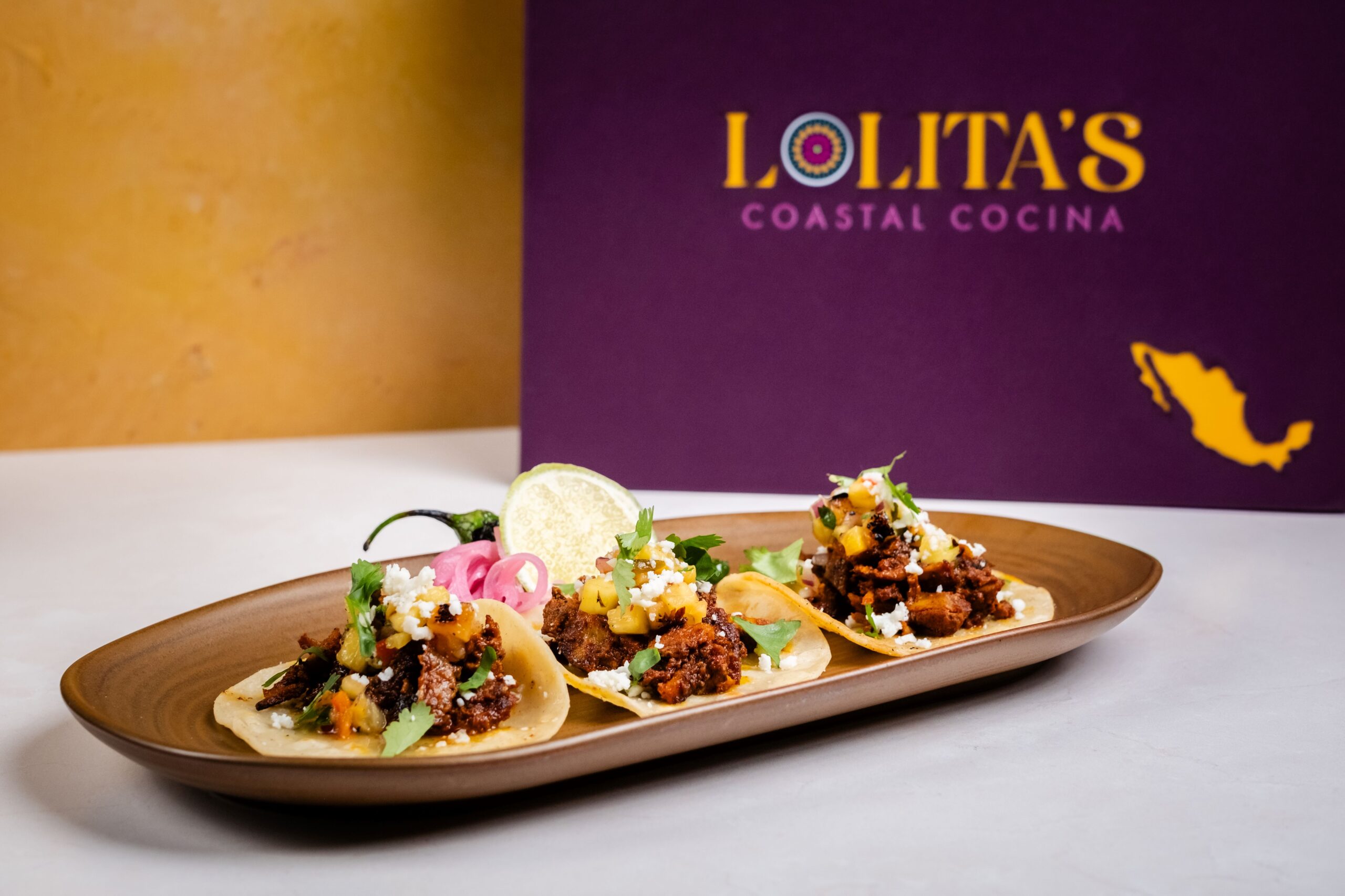 A plate of three tacos with various toppings, garnished with lime and herbs, is placed on a table. Behind it, theres a purple menu cover displaying Lolitas Coastal Cocina with a small map of Mexico.