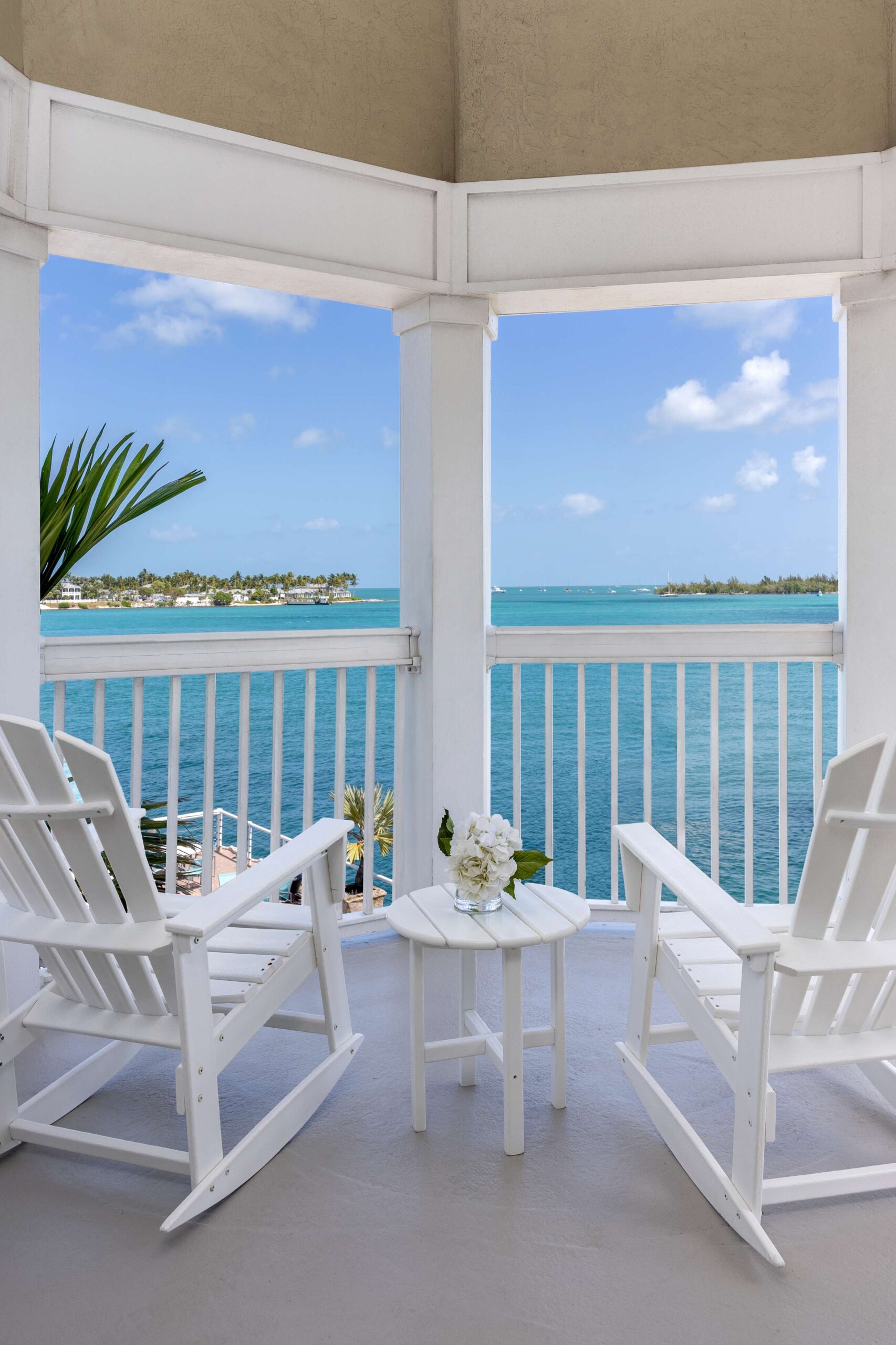 Two white rocking chairs and a small table with flowers sit on a balcony overlooking a calm blue ocean, with a view of distant land and a partly cloudy sky.