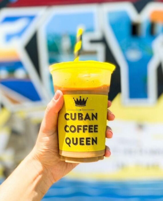 A hand holds a plastic cup with a yellow lid and straw, labeled Cuban Coffee Queen in black text, with a colorful, out-of-focus mural in the background.