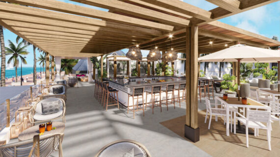 Open-air beachside bar and dining area with wooden pergola, modern bar stools, wicker chairs, white tables, and tropical plants. Ocean and palm trees are visible in the background under a sunny sky.