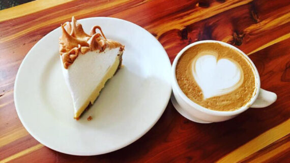 A slice of pie with a fluffy meringue topping sits on a white plate next to a cup of coffee with a heart-shaped latte art on a wooden table.