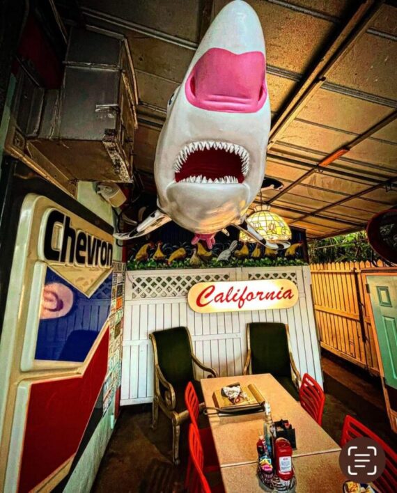A quirky indoor dining area with retro decor, including a Chevron sign, a California sign, and a large shark model hanging from the ceiling above a table set for four.